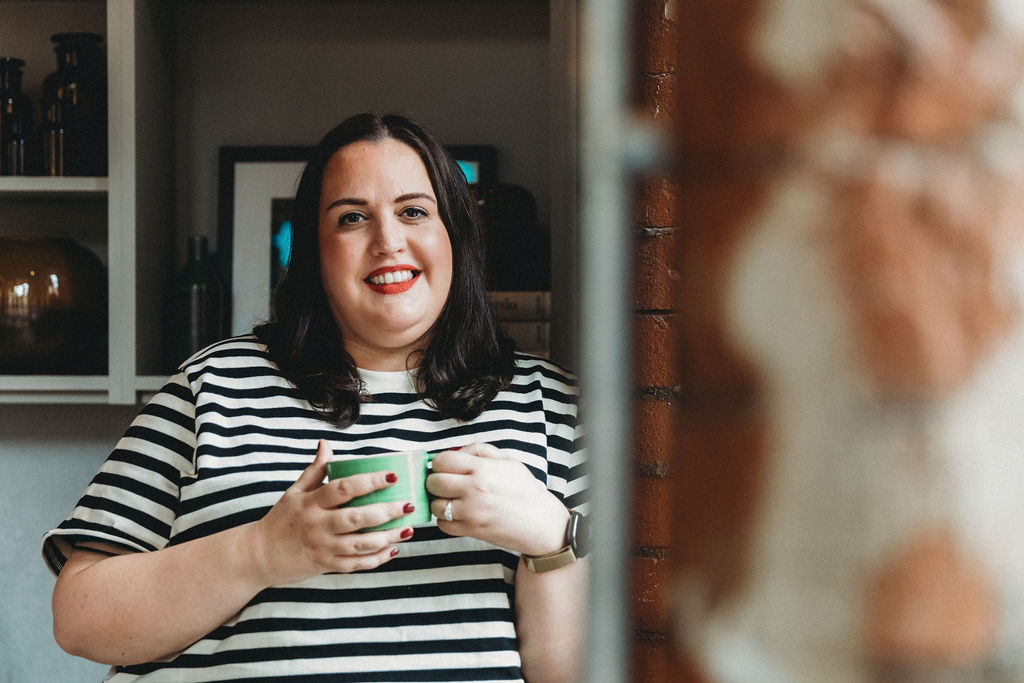 A photograph of Alice, a dark haired white woman wearing a black and cream stripe top, holding a green mug. Alice is smiling at the camera